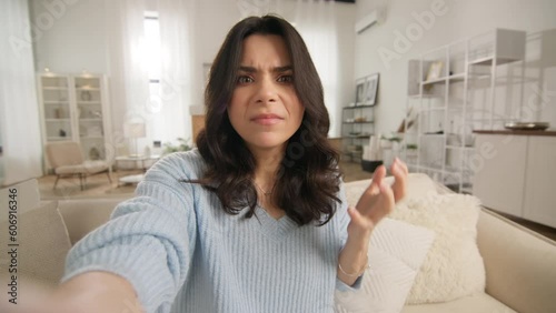 Portrait of concerned, uncertained and frustrated female with beautiful hair doing video chat in apartment. Puzzled young African American woman making selfie or video call while sitting on sofa home photo