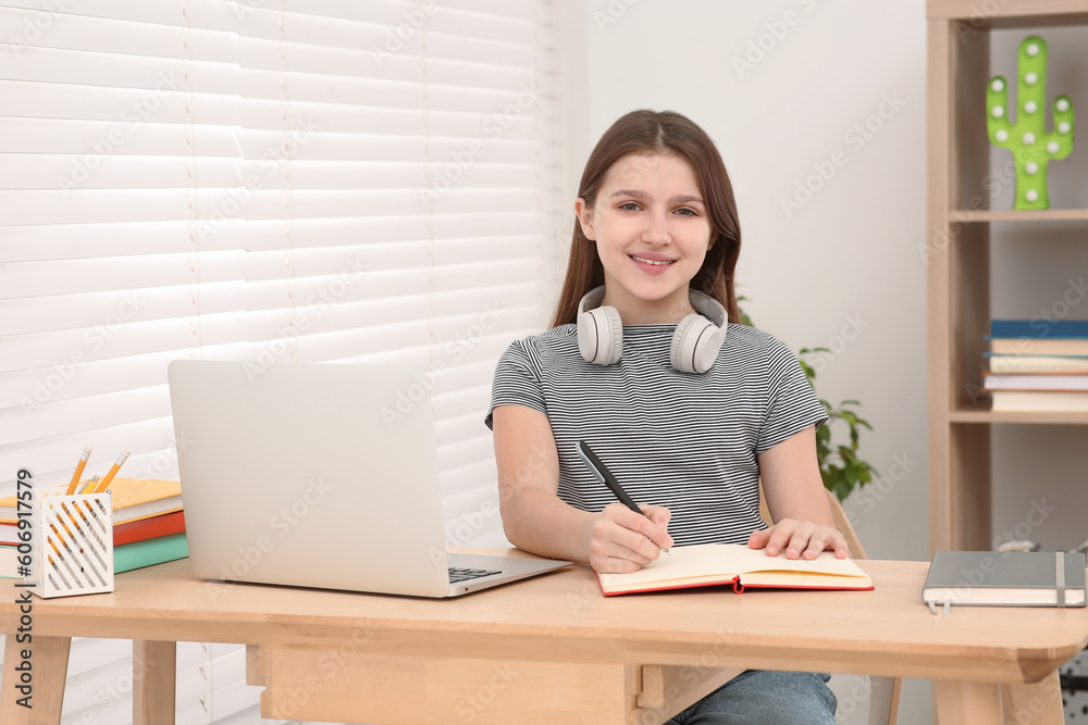 Cute girl with headphones writing in notepad near laptop at desk in room. Home workplace