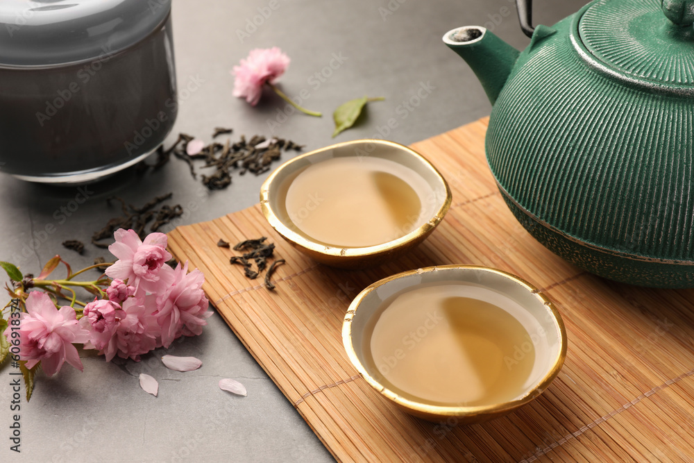 Traditional ceremony. Cups of brewed tea, teapot and sakura flowers on grey table, closeup - obrazy, fototapety, plakaty 