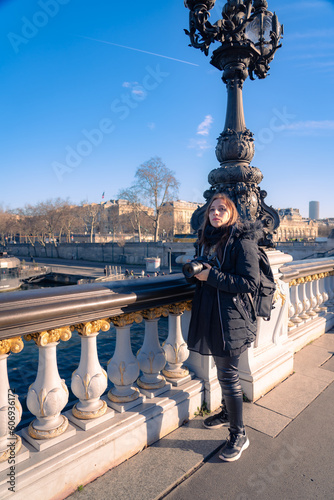 Full body of thoughtful and pensive young Latin woman traveler in warm outfit standing on Pont Alexandre bridge with photo camera and looking away during sightseeing trip in Paris  France