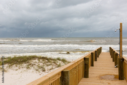 Wooden boardwalk with outdoor shower next to grassy sand dune on beach with brown and white surf waves under a cloudy sky before a storm