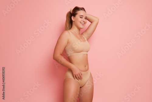 Pretty girl stands on pink background in ergonomic underwear of beige colour smiles and holds her hair with her hand  body comfort concept  copy space