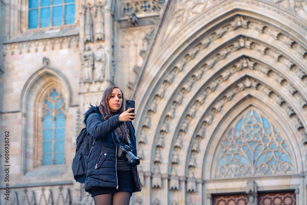 Calm young Latin woman tourist in warm clothes with backpack and camera taking selfie on smartphone while standing against ancient Barcelona Cathedral in Gothic Quarter Spain during sightseeing tour