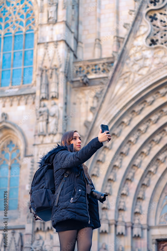 Calm young Latin woman tourist in warm clothes with backpack and camera taking selfie on smartphone while standing against ancient Barcelona Cathedral in Gothic Quarter Spain during sightseeing tour
