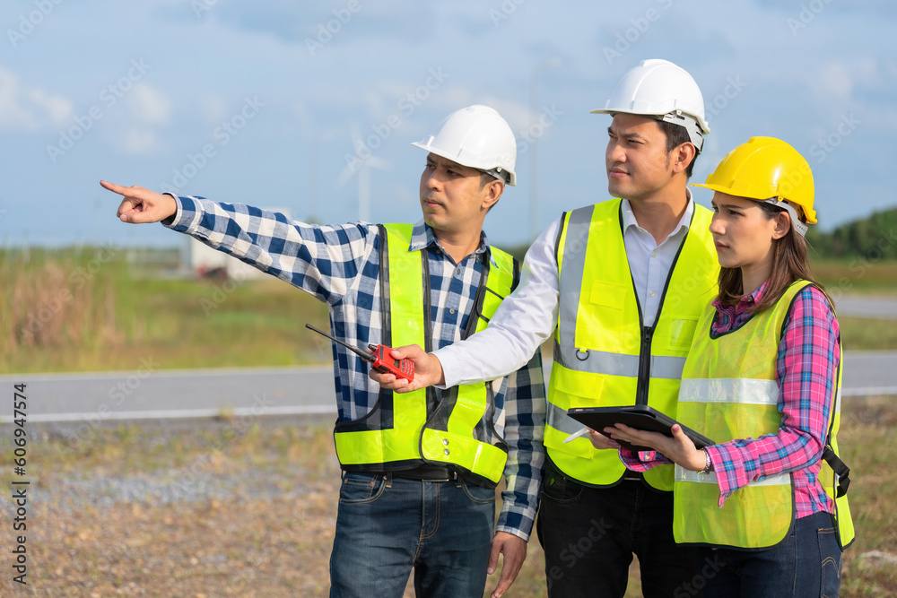 A team of 3 male and female engineers or architects working on a project meeting near an industrial turbine. pointing to the target ahead wearing a helmet and vest holding a tablet A team of 3 male and female engineers or architects working on a project meeting near an industrial turbine. pointing to the target ahead wearing a helmet and vest holding a tablet