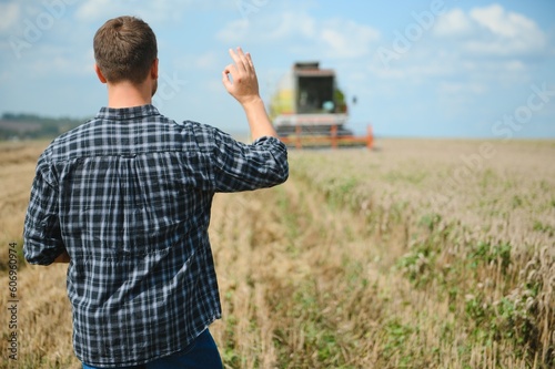 Happy farmer proudly standing in a field. Combine harvester driver going to crop rich wheat harvest. Agronomist wearing flannel shirt, looking at camera on a farmland