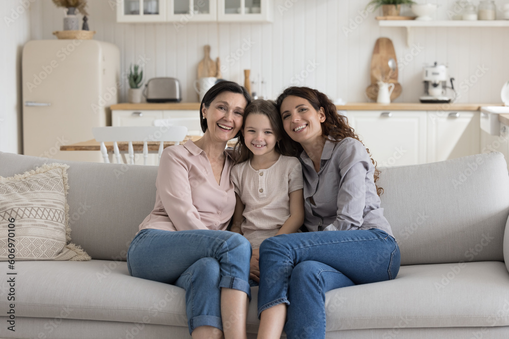 Cheerful child girl, mother and grandma sitting close on home couch, hugging with love, affection, enjoying closeness, bonding, close relationship, laughing, smiling. Portrait of three generations