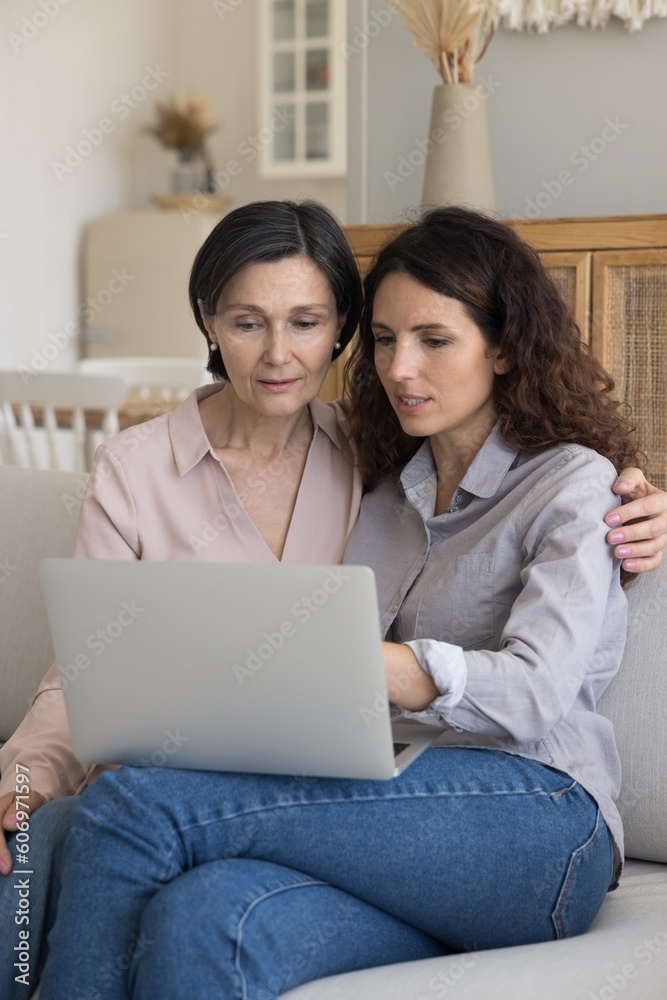 Adult daughter woman showing financial banking payment application on laptop to mature mom, helping with shopping, online technology, using computer for Internet communication