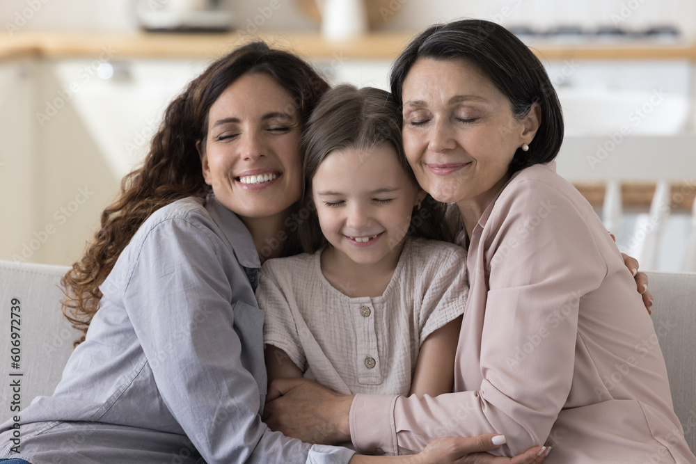 Positive loving mom and grandma hugging preschool kid, cuddling, tightening, tickling cute little girl, smiling with closed eyes, enjoying closeness, childcare, family relationships Positive loving mom and grandma hugging preschool kid, cuddling, tightening, tickling cute little girl, smiling with closed eyes, enjoying closeness, childcare, family relationships