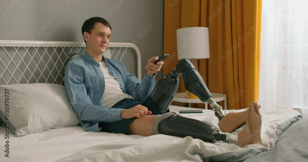 lonely disabled caucasian man with TV controller enjoying movie indoors. Nice young handicapped man with prosthesis watching television, switching between channels, alone at home, in casual wear lonely disabled caucasian man with TV controller enjoying movie indoors. Nice young handicapped man with prosthesis watching television, switching between channels, alone at home, in casual wear