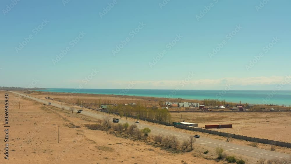 Aerial view of busy highway with trucks and cars along picturesque lake and small village. Road trip, Kyrgyzstan, Central Asia. Aerial view of busy highway with trucks and cars along picturesque lake and small village. Road trip, Kyrgyzstan, Central Asia.