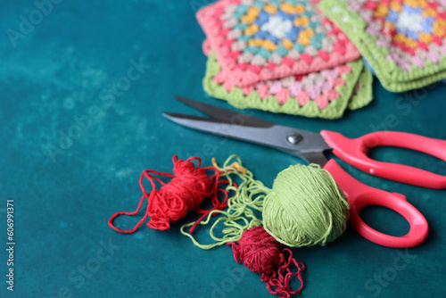 Fototapeta Naklejka Na Ścianę i Meble -  Red scissors, cotton yarn balls, crochet hook and granny squares on a blue desk. Messy working place. Crochet geometric shape. 
