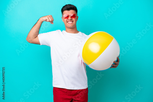 Young caucasian man holding a beach ball isolated on blue background doing strong gesture