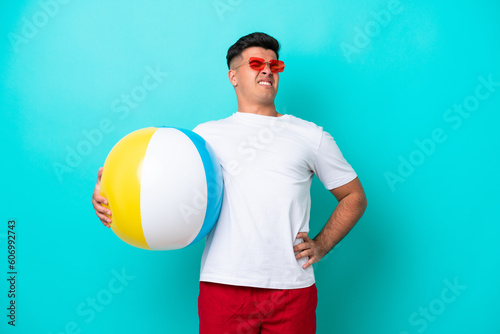 Young caucasian man holding a beach ball isolated on blue background suffering from backache for having made an effort