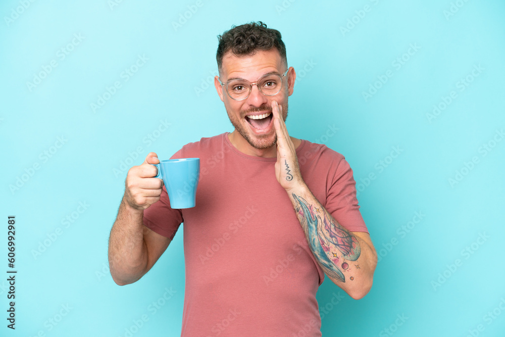 Young caucasian man holding cup of coffee isolated on blue background shouting with mouth wide open Young caucasian man holding cup of coffee isolated on blue background shouting with mouth wide open