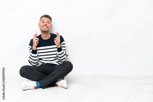 Young man sitting on the floor isolated on white background with fingers crossing