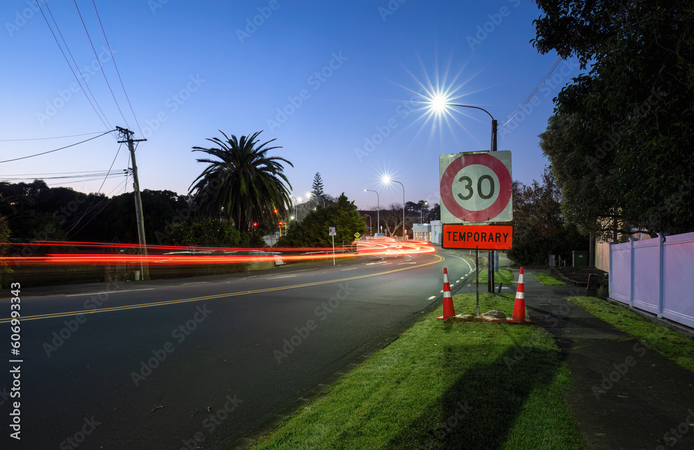 Temporary 30km speed limit sign with traffic cones by the roadside. Car light trails on the road intersection. Auckland.