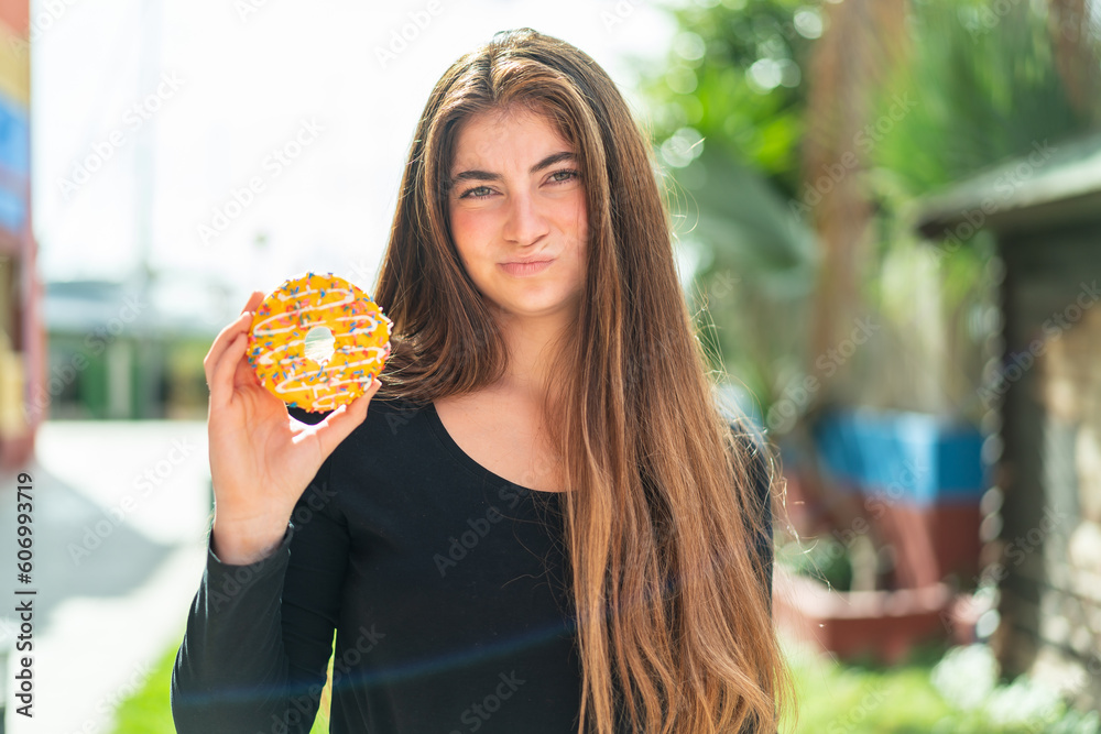 Young pretty caucasian woman holding a donut at outdoors with sad expression