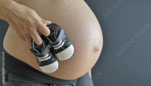 A young pregnant woman holding  baby shoes, against the backdrop of her pregnant belly.