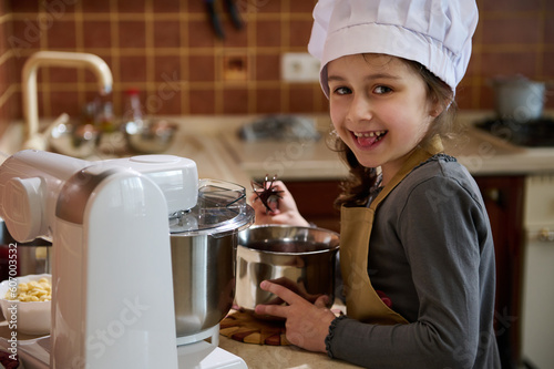 Mischievous child girl 5 years old, tastes melted Belgian chocolate, standing at the kitchen countertop, smiling slyly at the camera. Little kid pastry chef dressed in white chef's hat and beige apron photo