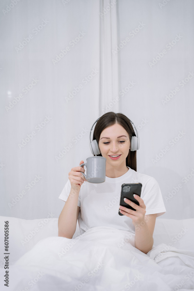 Smiling girl relaxing at home, she is playing music using smartphone wearing white headphones and enjoys a milk in the morning. Smiling girl relaxing at home, she is playing music using smartphone wearing white headphones and enjoys a milk in the morning.