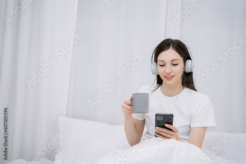 Smiling girl relaxing at home, she is playing music using smartphone wearing white headphones and enjoys a milk in the morning.