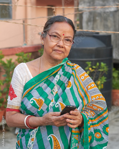 An aged Bengali woman (Indian ethnicity) in sari, holding a basic phone in hand. Photo taken on a building rooftop, in Kolkata, West Bengal.