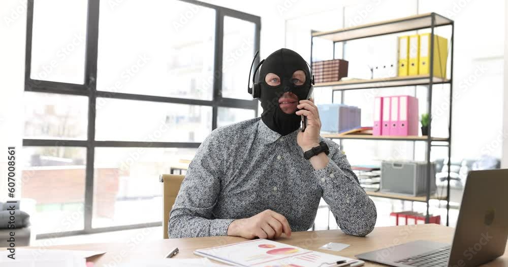 Fake manager in mask of robber talks to client sitting at workplace in company premise. Male scammer in headphones communicates cheating customer via call