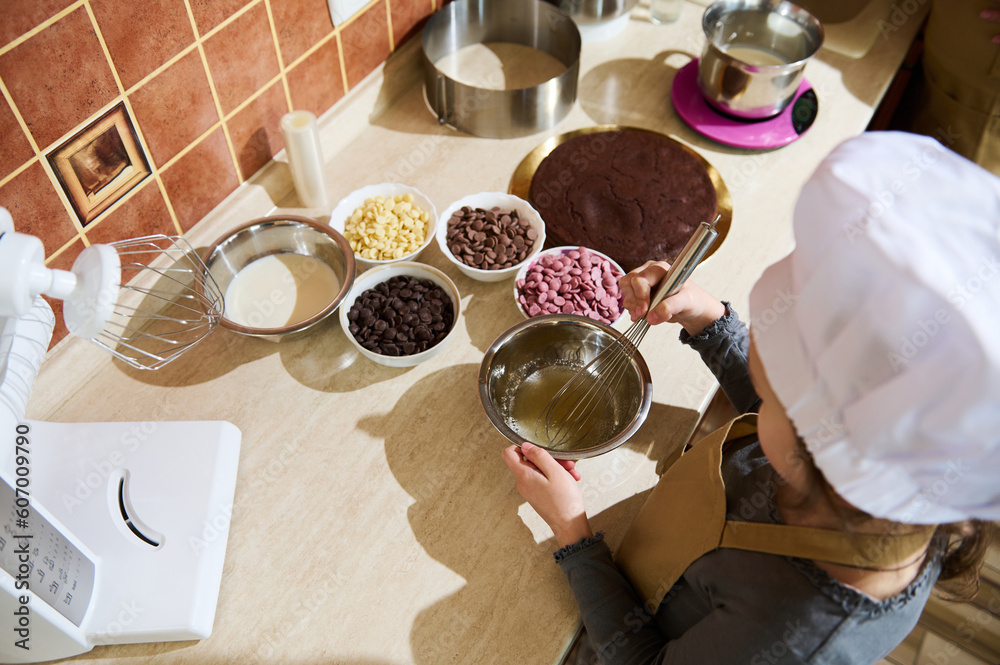 Overhead view of a cute child, little baker confectioner using whisk, mixing sugar with syrup in a metal bowl, standing by kitchen countertop with various ingredients and drops of belgian chocolate Overhead view of a cute child, little baker confectioner using whisk, mixing sugar with syrup in a metal bowl, standing by kitchen countertop with various ingredients and drops of belgian chocolate