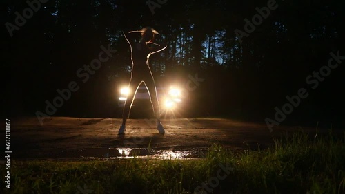 A young slender girl dances a modern dance against the background of car headlights in a night forest