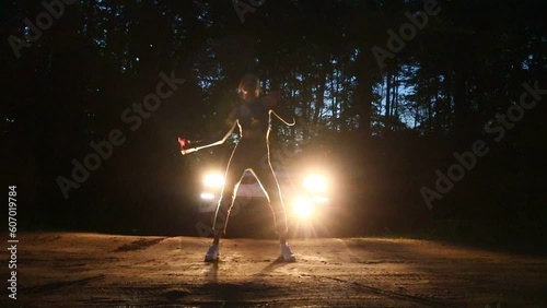 A young slender girl dances a modern dance against the background of car headlights in a night forest