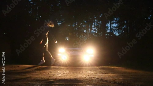A young slender girl dances a modern dance against the background of car headlights in a night forest
