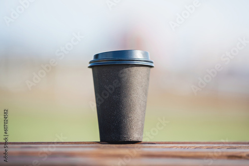 Paper coffee cup with empty space for advertising information stands on wooden terrace table against blurred nature background.
