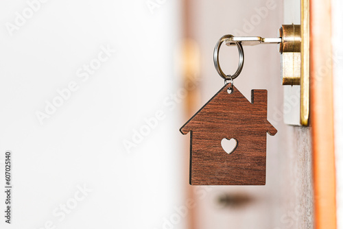 Wooden key chain in the shape of a house with a heart-shaped hole in the center hanging from a key inserted in the lock of a front door of a house.