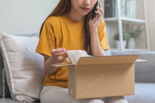 Angry bad, complaint asian young woman opening carton box, received online shopping parcel wrong product order from retail store, using mobile phone talking with support shop want to return package. photo