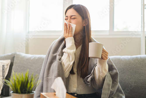 Sick, hurt or pain asian young woman have a fever, flu sore throat, drinking a mug of warm water, use tissues paper sneezing nose, runny while sitting rest on sofa at home. Health care on virus person
