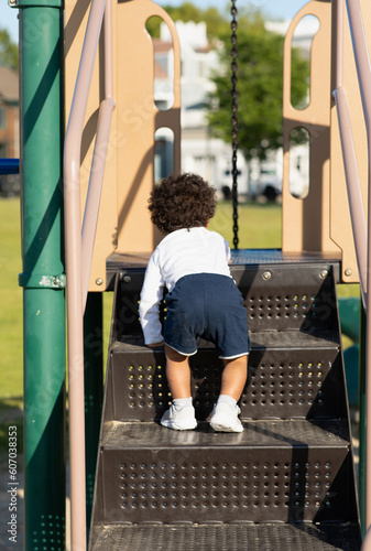 A young boy is climbing up large stairs on a playground. A toddler is learning to walk and step up. The baby is working on gross motor development, reaching toddler milestones by practicing outside. 