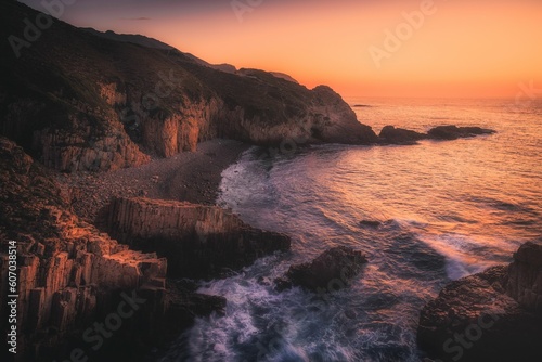Waves crashing the cliffs of a beach during a sunset © Tang Pan/Wirestock Creators
