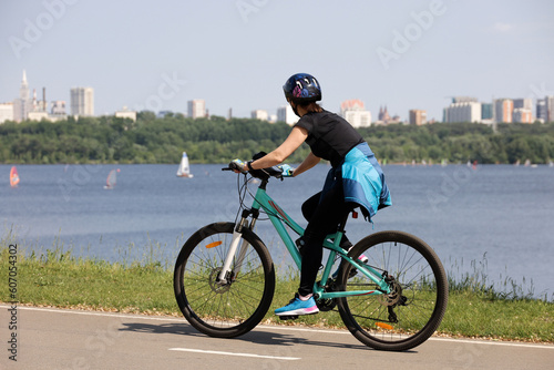 Girl in sportswear and helmet riding bicycle on river and city background. Cycling and summer leisure