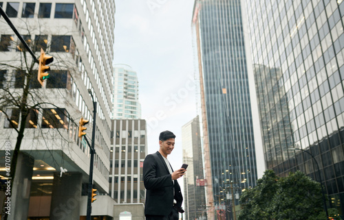 Young happy Asian businessman executive, Japanese business man investor standing in big city street with downtown buildings using cell phone looking at smartphone checking financial apps on mobile.