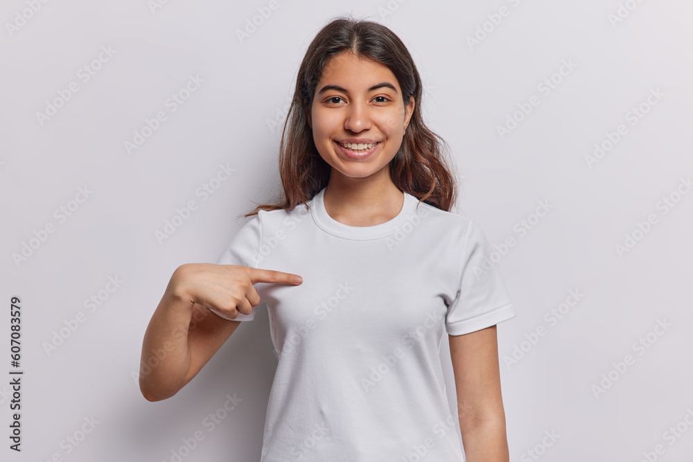 Smiling dark haired young woman from Iran demonstrates her new white casual t shirt being glad after making purchase indicates at blank copy space for your designing content or logo poses indoor Smiling dark haired young woman from Iran demonstrates her new white casual t shirt being glad after making purchase indicates at blank copy space for your designing content or logo poses indoor