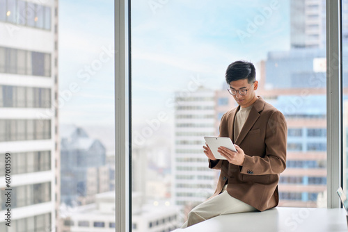 Busy young adult Asian business man professional manager, company executive using digital tablet fintech smart tech device in corporate modern office with big window, copy space, authentic shot.