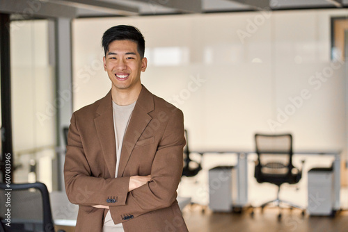 Young happy Asian business man looking at camera standing in office. Smiling confident professional Japanese businessman executive, successful software engineer or entrepreneur wearing suit, portrait.