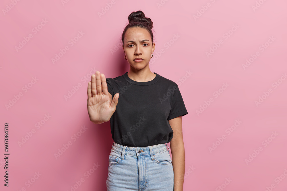 Serious Latin woman shows stop gesture with palm asserts boundaries sends message of resistance has strict expression wears black t shirt and jeans poses against pink background. Self empowerment - obrazy, fototapety, plakaty 