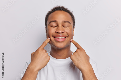 Pleased short haired man points at toothy smile keeps eyes closed has cheerful expression dressed in casual t shirt isolated over white background. Genuine happinness positive human emotions concept photo