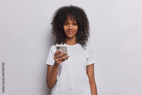 Pleased curly haired woman with satisfied expression holds mobile phone types message or scrolls news in internet dressed in casual t shirt isolated on white background. People and technology concept