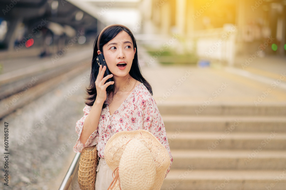 Fototapeta premium Asian young woman traveler with weaving hat using a mobile phone beside railway train station in Bangkok. Journey trip lifestyle, world travel explorer or Asia summer tourism concept.