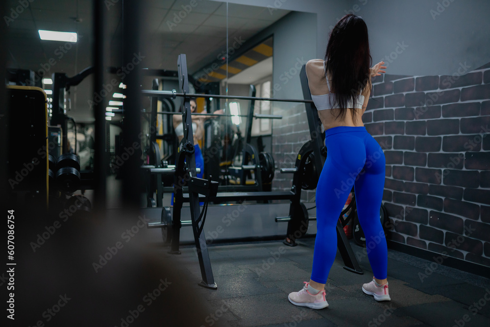 A sports girl in blue leggings and a sexy body warms up in front of a mirror at a workout in a gym with exercise equipment	