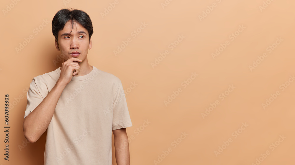 Thoughtful Asian man keeps hand on chin concentrated aside pensively ponders on complicated choice wears casual t shirt isolated over brown background copy space for your promotional content Thoughtful Asian man keeps hand on chin concentrated aside pensively ponders on complicated choice wears casual t shirt isolated over brown background copy space for your promotional content