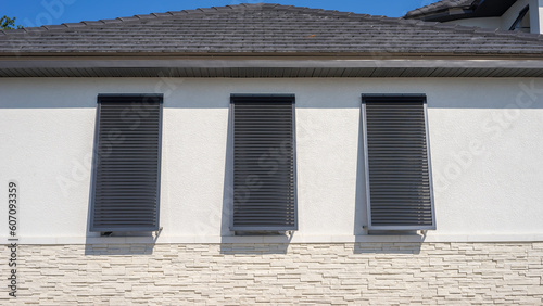 Three storm shutters, hinged at the top, over three tall windows on an upscale suburban house under construction on a sunny morning in southwest Florida